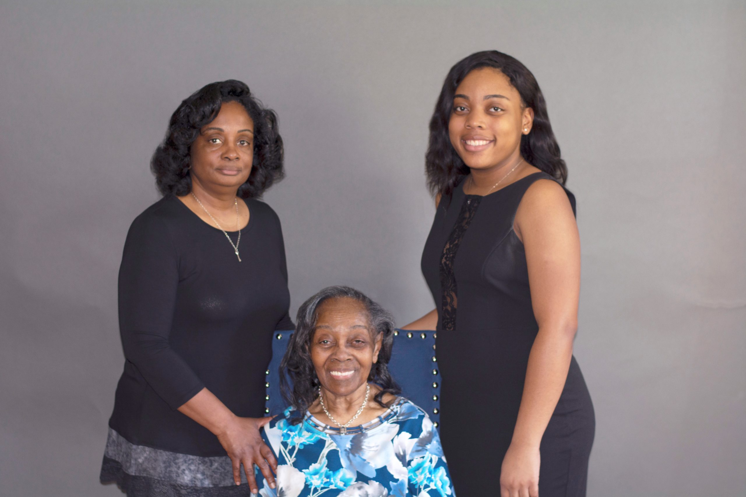 2 women standing up on each side of the grandmother whom is sitting down in a chair