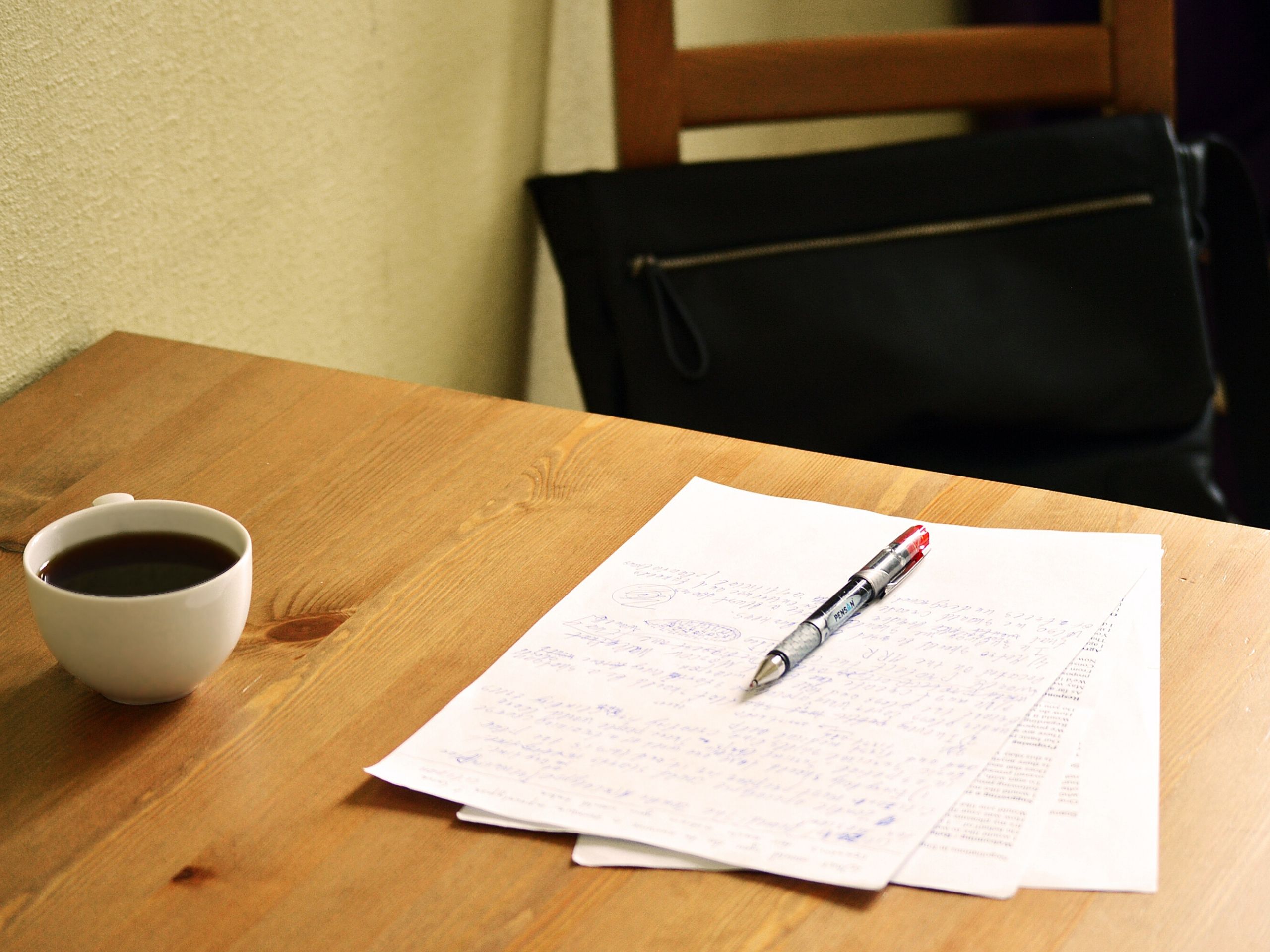 Pen and paper on a table with a small cup of coffee and leather bag on the chair in the background