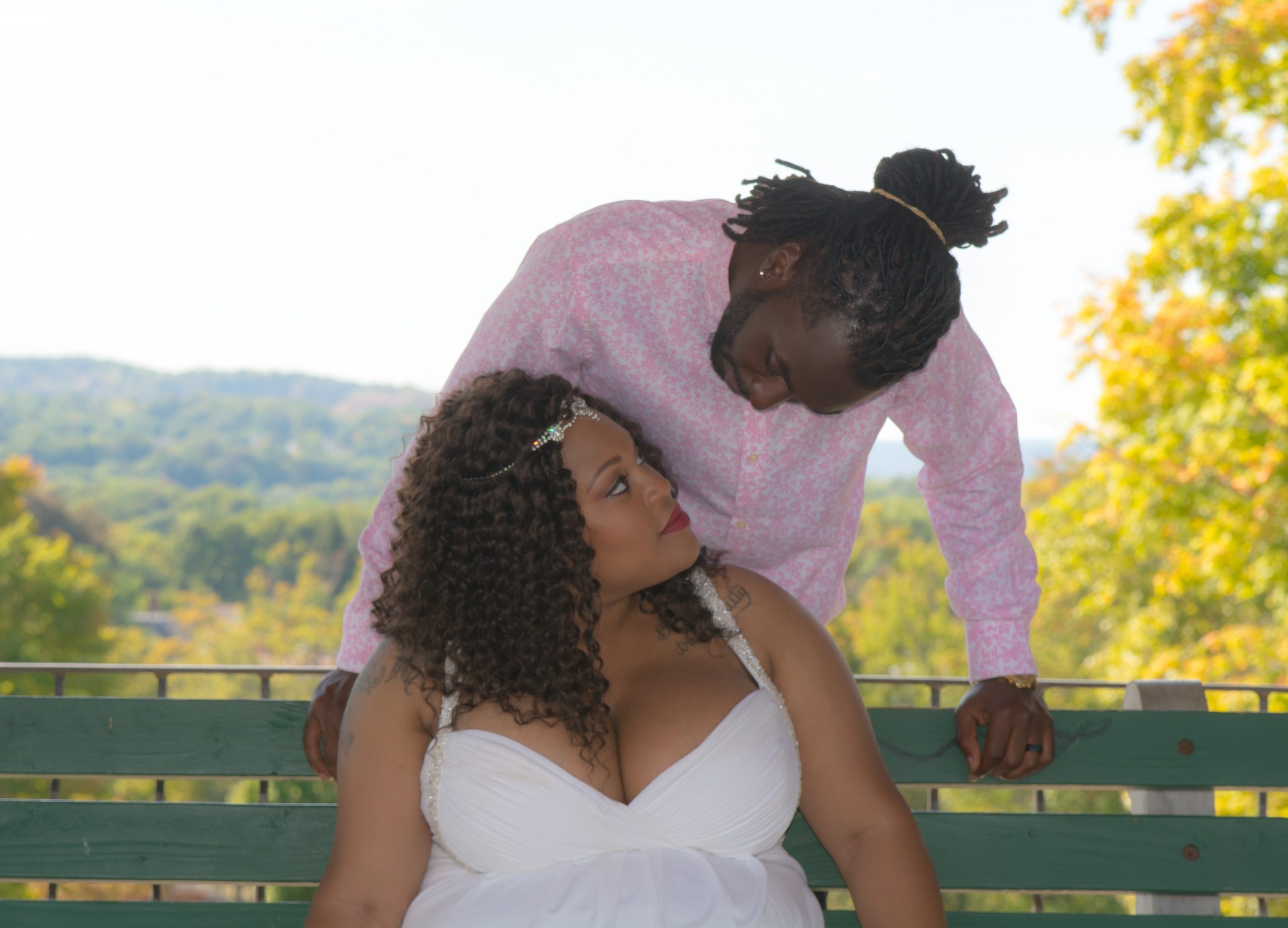 Bride sitting on a park bench looking up at groom while he is standing behind her looking back down at her