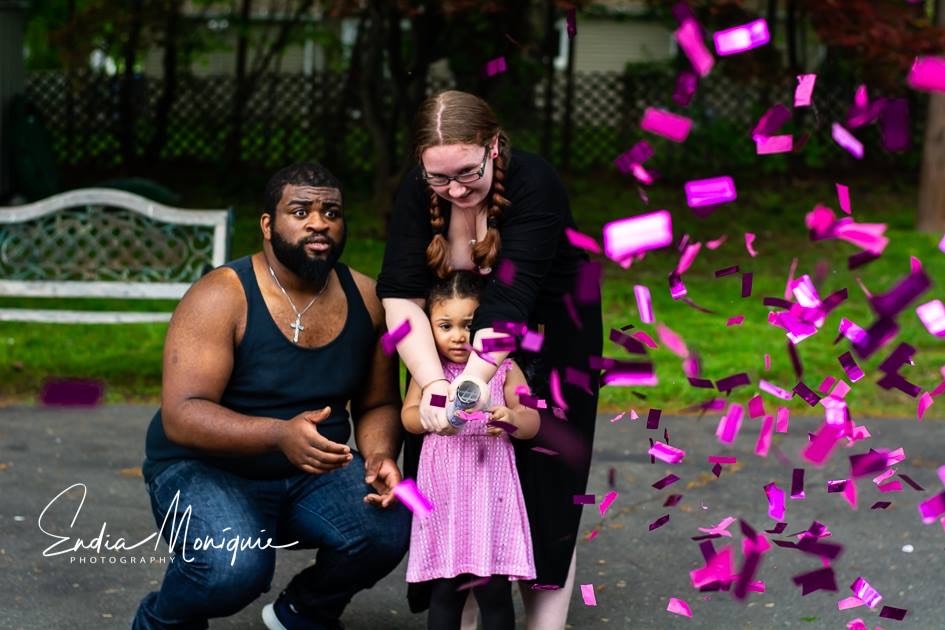 Mother helping her daughter set off the confetti cannon while the dad looks on shocked that it is a girl